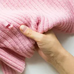 Dwie dłonie trzymają różową, dzianinową bluzkę z widoczną dziurą. Pokazano, jak załatać dziurę w bluzce.