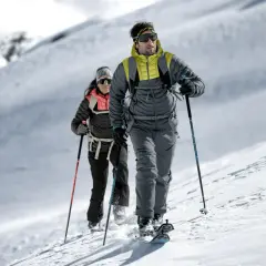Dwóch narciarzy skitourowych w zimowych kurtkach, z kijkami i nartami, przemierza ośnieżone zbocze.