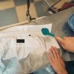 Ręce czyszczą białą kurtkę z plam, używając szczoteczki i wody z mydłem. Dowiedz się, jak zmyć farbę z kurtki.
