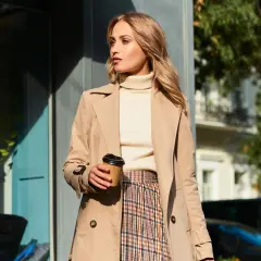 Elegancka kobieta w beżowym płaszczu, kremowym golfie i kraciastej spódnicy, z kubkiem kawy w dłoni. Inspiracja na stylizacje do beżowego płaszcza.