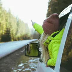 Dziewczyna w jaskrawozielonej kurtce i bordowej czapce wystawia rękę przez okno samochodu, wskazując palcem. Idealne dodatki do zielonej kurtki na podróż!