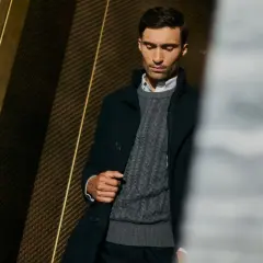 Mężczyzna w eleganckim swetrze jak marynarka, z warkoczowym splotem, w ciemnej, stylowej stylizacji.
