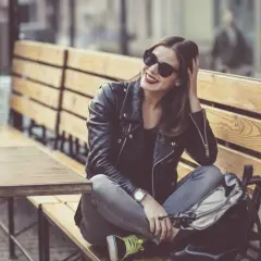 Dziewczyna w skórzanej kurtce i okularach przeciwsłonecznych, z uśmiechem na twarzy, siedzi na ławce w kawiarni. Jej styl jest zdecydowanie rockowy.