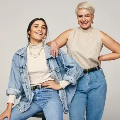 Dwie uśmiechnięte kobiety w jeansach. Jedna w beżowym golfie i jeansowej kurtce, druga w beżowej bluzce bez rękawów. Inspiracja: jaka bluzka do jeansów.