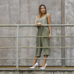 Kobieta w zielonym kombinezonie i białych szpilkach stoi przy metalowej balustradzie na tle betonowej ściany.