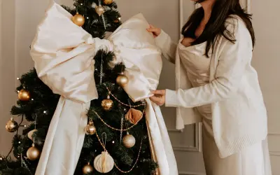 Elegancka stylizacja na święta bożego narodzenia: kobieta w satynowej sukience i swetrze poprawia dużą, kremową kokardę na choince ozdobionej złotymi bombkami.