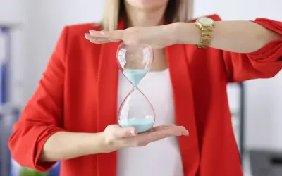 Kobieta w czerwonej marynarce trzyma klepsydrę z niebieskim piaskiem, symbolizując upływ czasu.