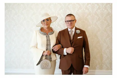 Elegancka mama pana młodego w kremowym kostiumie i kapeluszu, obok syna w brązowym garniturze.