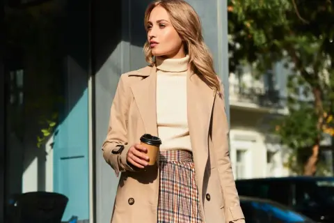 Elegancka kobieta w beżowym płaszczu, kremowym golfie i kraciastej spódnicy, z kubkiem kawy w dłoni. Inspiracja na stylizacje do beżowego płaszcza.