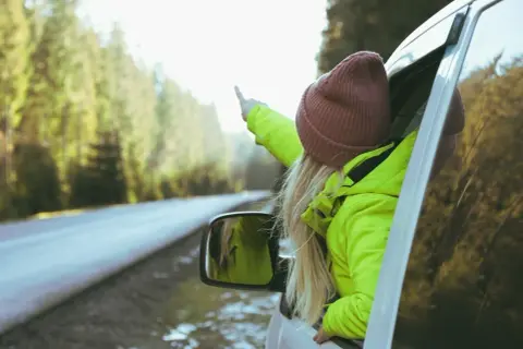 Dziewczyna w jaskrawozielonej kurtce i bordowej czapce wystawia rękę przez okno samochodu, wskazując palcem. Idealne dodatki do zielonej kurtki na podróż!