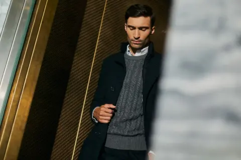 Mężczyzna w eleganckim swetrze jak marynarka, z warkoczowym splotem, w ciemnej, stylowej stylizacji.