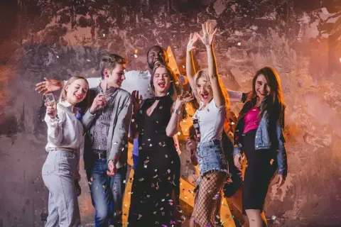 Grupa młodych ludzi świętuje, sypią się konfetti. Inspiracja, jak się ubrać do klubu: sukienki, jeansy, marynarki.