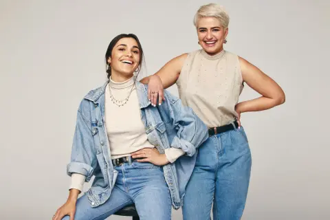 Dwie uśmiechnięte kobiety w jeansach. Jedna w beżowym golfie i jeansowej kurtce, druga w beżowej bluzce bez rękawów. Inspiracja: jaka bluzka do jeansów.