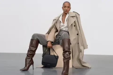 Eleganckie stylizacje z koszulą oversize: modelka w beżowym prochowcu, szarych spodniach, brązowych kozakach i czarnej torebce.