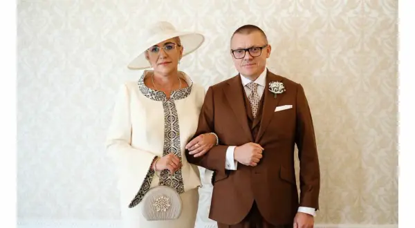 Elegancka mama pana młodego w kremowym kostiumie i kapeluszu, obok syna w brązowym garniturze.