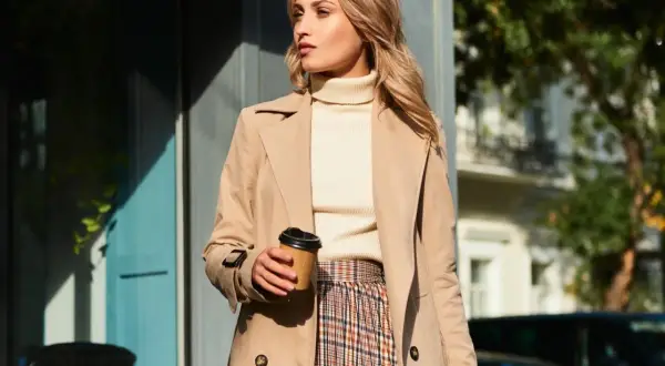 Elegancka kobieta w beżowym płaszczu, kremowym golfie i kraciastej spódnicy, z kubkiem kawy w dłoni. Inspiracja na stylizacje do beżowego płaszcza.