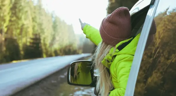 Dziewczyna w jaskrawozielonej kurtce i bordowej czapce wystawia rękę przez okno samochodu, wskazując palcem. Idealne dodatki do zielonej kurtki na podróż!