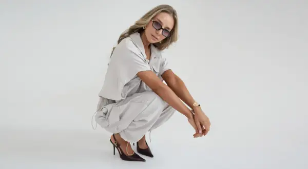 Kobieta w beżowym, luźnym komplecie i szpilkach prezentuje modne stylizacje damskie. Okulary i złota bransoletka dodają szyku.