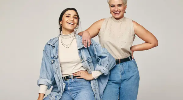 Dwie uśmiechnięte kobiety w jeansach. Jedna w beżowym golfie i jeansowej kurtce, druga w beżowej bluzce bez rękawów. Inspiracja: jaka bluzka do jeansów.