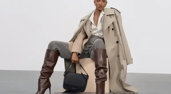 Eleganckie stylizacje z koszulą oversize: modelka w beżowym prochowcu, szarych spodniach, brązowych kozakach i czarnej torebce.