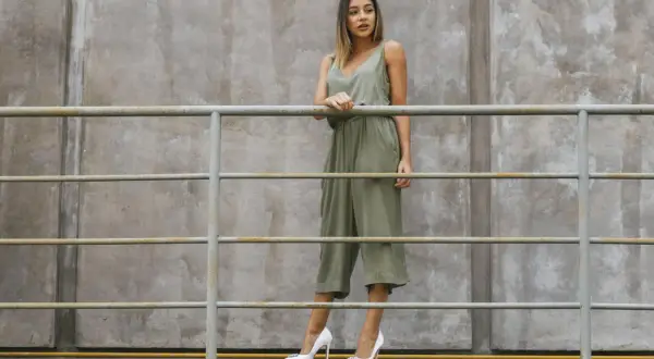 Kobieta w zielonym kombinezonie i białych szpilkach stoi przy metalowej balustradzie na tle betonowej ściany.