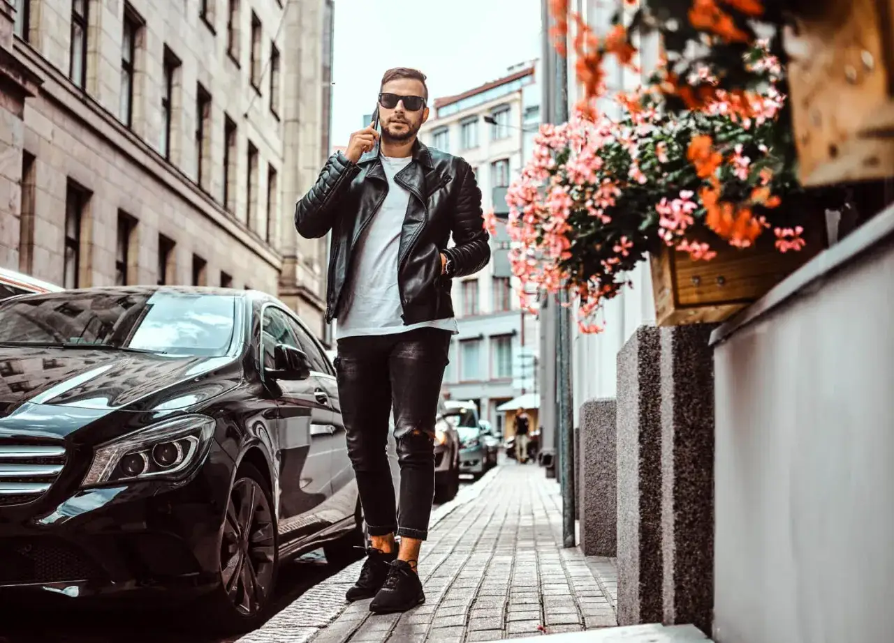 Elegancki mężczyzna w skórzanej kurtce i czarnych sneakersach, idealne jakie buty do skórzanej kurtki facet. Spaceruje ulicą obok czarnego samochodu.