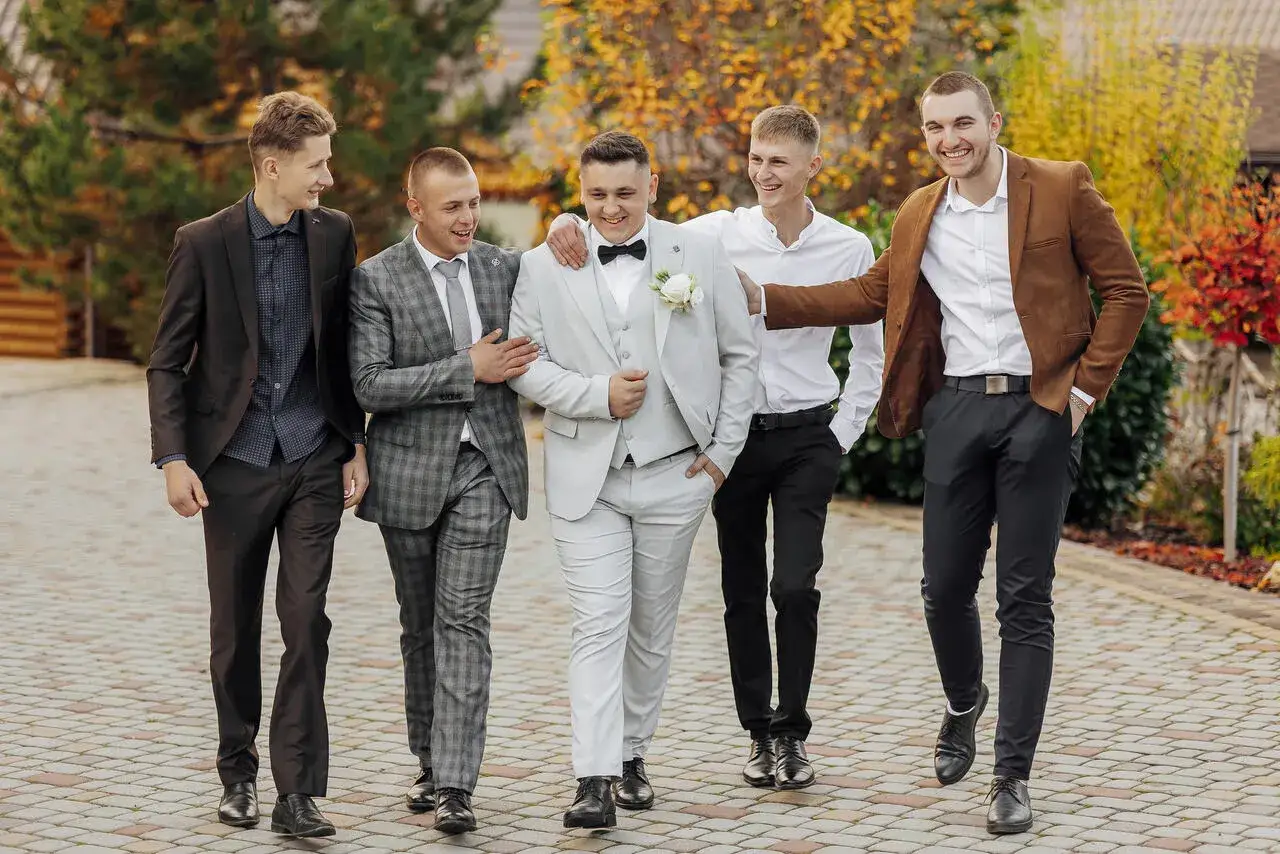 Pięciu młodych mężczyzn w eleganckich strojach formalnych, śmiejąc się, idzie razem po brukowanej ścieżce.