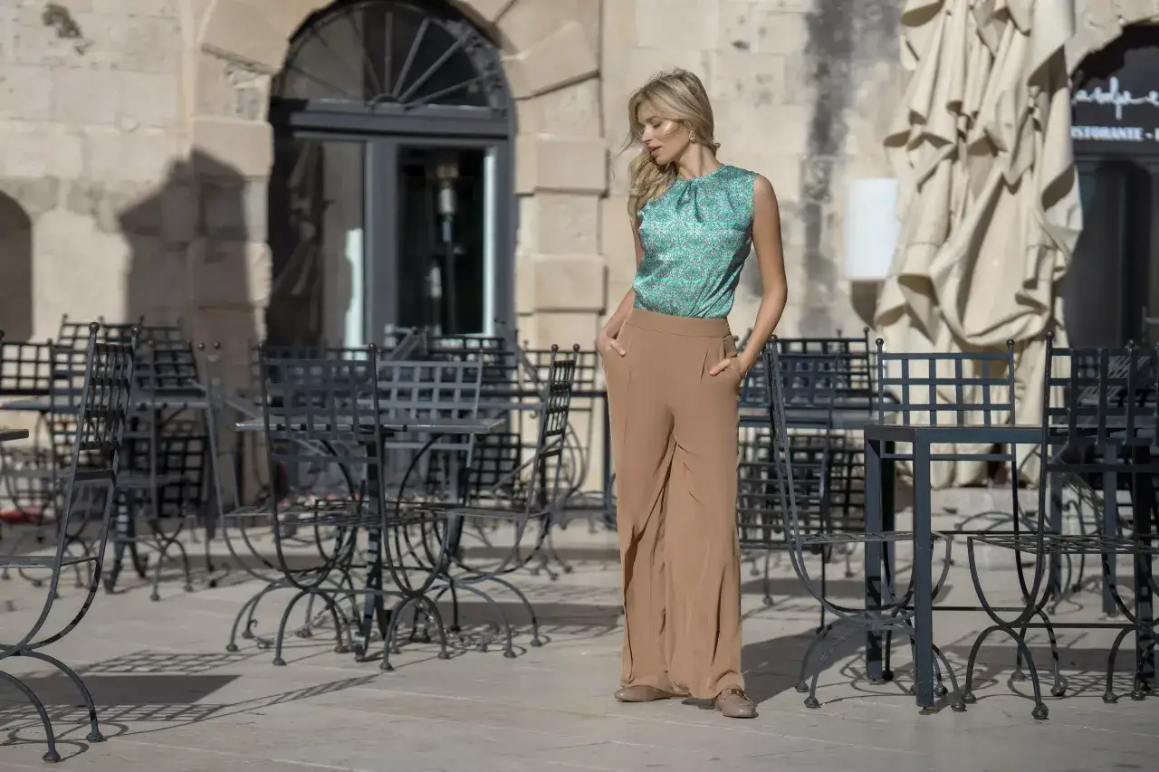 Elegancka stylizacja na egzamin: zielona bluzka w delikatny wzór i beżowe, szerokie spodnie. Kobieta stoi na zewnątrz, przed kawiarnią.
