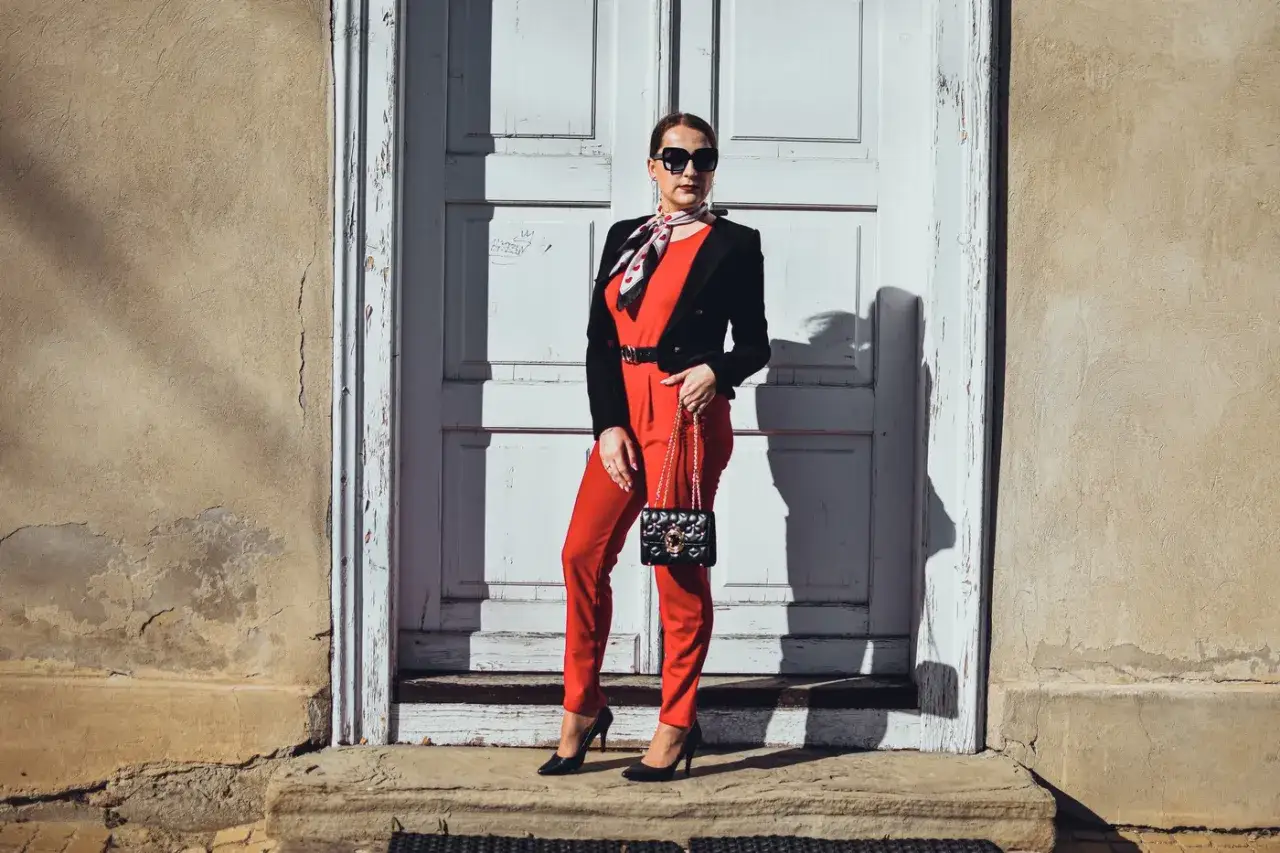Elegancka kobieta w czerwonym kombinezonie i czarnym żakiecie, zastanawia się, jakie buty do czerwonego kombinezonu będą najlepsze. Czółenka idealnie pasują.