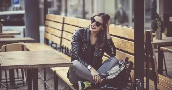 Dziewczyna w skórzanej kurtce i okularach przeciwsłonecznych, z uśmiechem na twarzy, siedzi na ławce w kawiarni. Jej styl jest zdecydowanie rockowy.